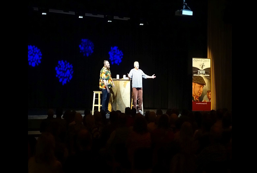 Bühne verdunkelt mit 2 Männern, die Comedy aufführen, rechts steht ein Plakat mit Schrift Pfefferle und Zipperle, woisch no?