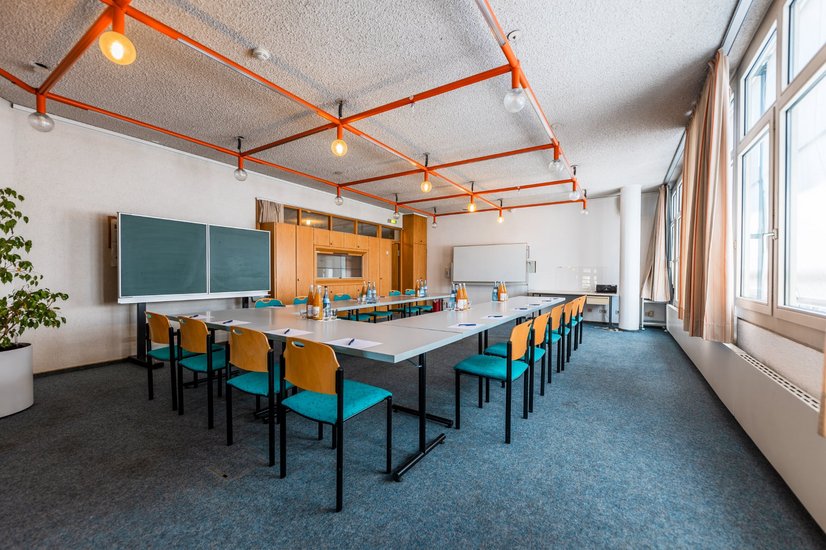 Seminarraum mit weißen Tischen im Rechteck und Stühlen, links steht große Schreibtafel